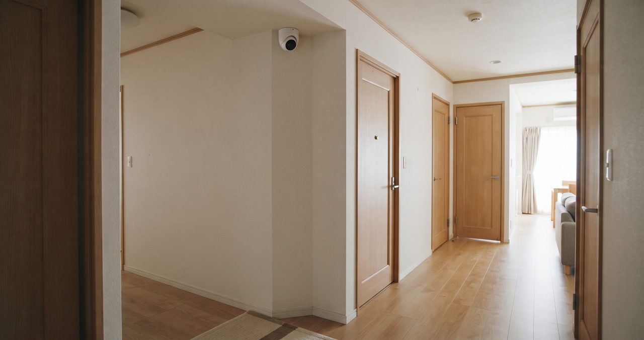 住宅の廊下の角に設置された見守りカメラが複数の部屋のドアを見渡している様子、2LDKの間取りで動線をカバーする設置例